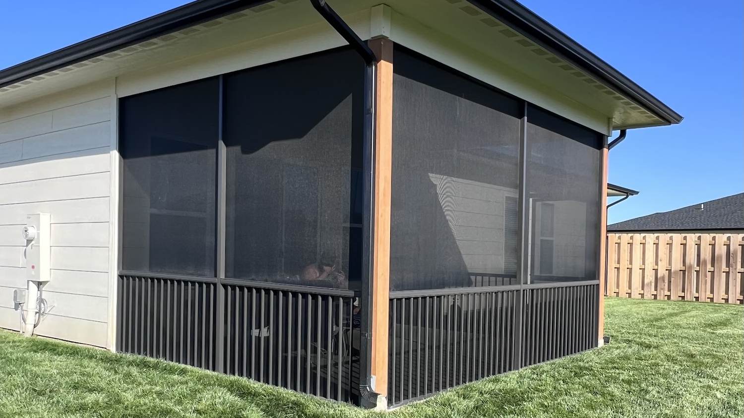 A black aluminum screen room built onto the back corner of a tan house, with a cedar post at the corner and a black rail along the base, above a green lawn.