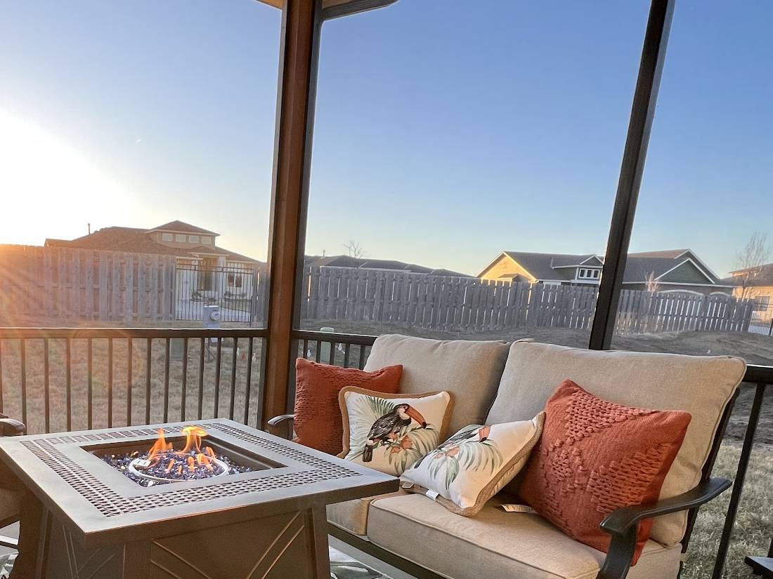A covered deck at sunset with a black metal rail, a square fire table with a lit flame, and a cushioned sofa with orange and toucan-print pillows.