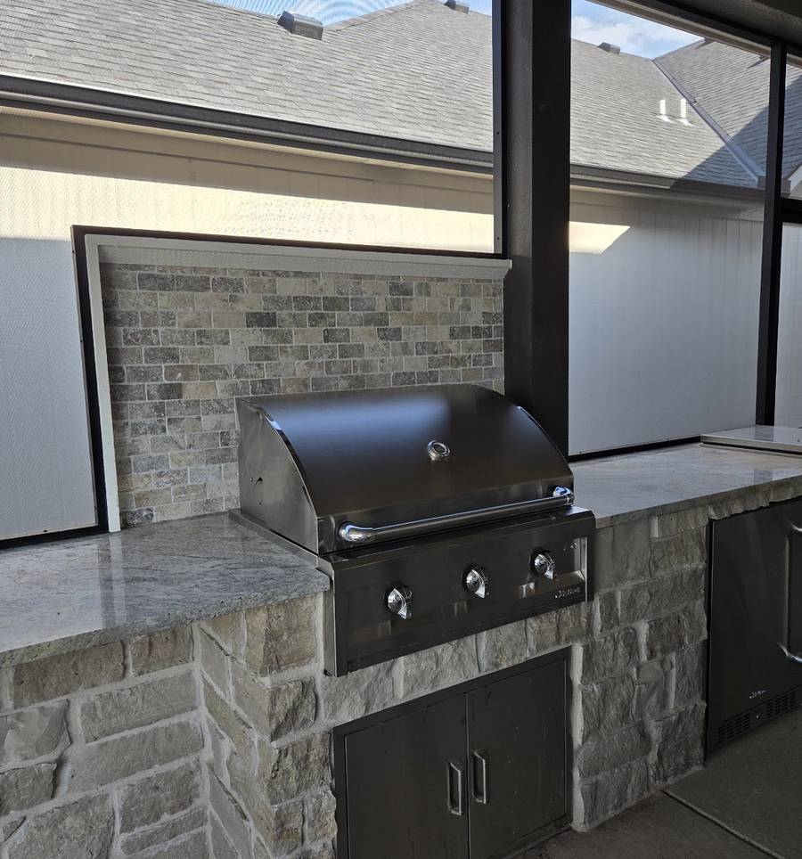 A stainless built-in gas grill set into a stone-faced counter on a covered patio, with a house in the background.