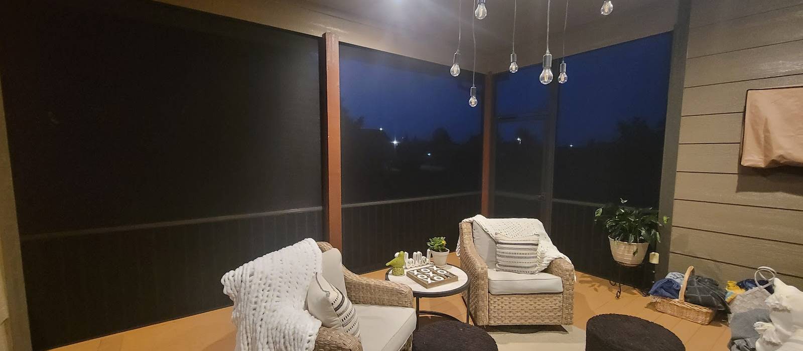 The inside of a dark screen room at dusk, with wicker chairs, a table and string lights above a wood-look floor.